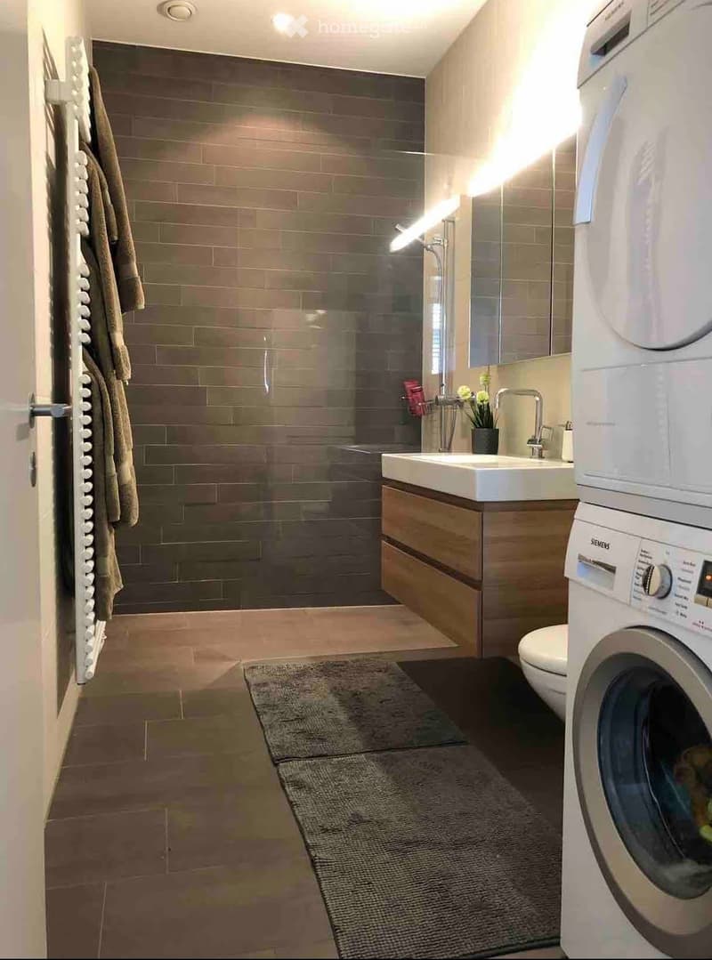 Modern bathroom with a glass-enclosed shower, a white sink with a wooden cabinet, a washing machine, and a towel rack with towels.