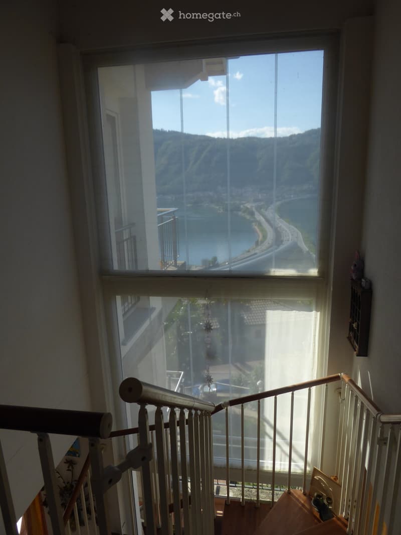 The image shows a room with large windows that provide a panoramic view of a mountainous landscape and a winding road below. The room appears to be on an upper floor, with a wooden railing visible in the foreground.