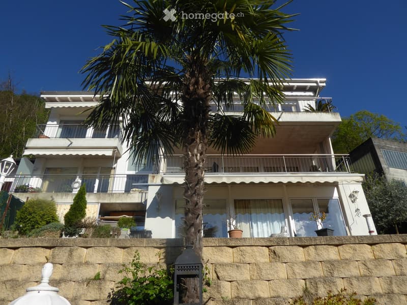 2-story white building with balconies, surrounded by palm trees and a stone wall