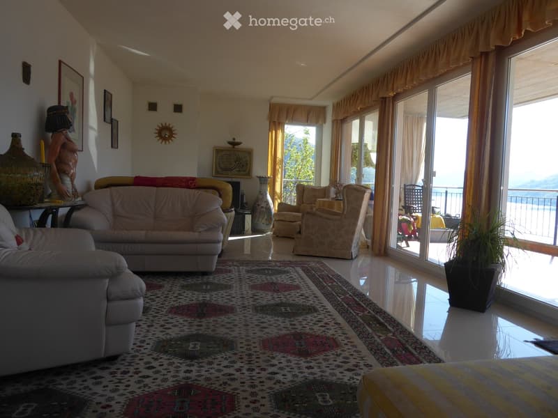 Spacious living room with large windows and sliding glass doors leading to a balcony with a scenic view. The room is furnished with a beige sofa, armchairs, and a patterned area rug. Decorative elements such as artwork, a sun sculpture, and potted plants
