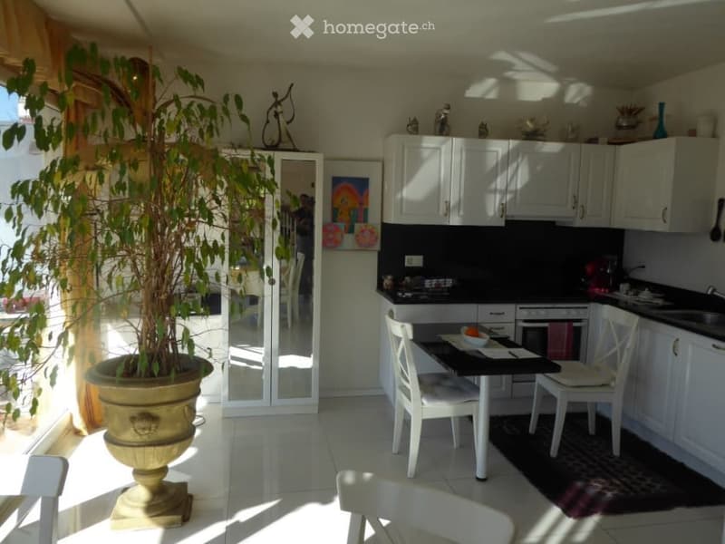 Bright and spacious kitchen with white cabinets, black countertops, and a dining table. There is a large potted plant in the corner, and artwork on the walls. The kitchen opens up to a balcony or terrace area.