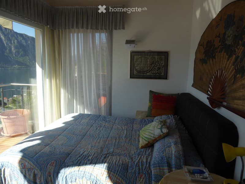 Spacious bedroom with large windows overlooking a scenic lake or mountain view, patterned bedding, decorative pillows, and a framed artwork on the wall