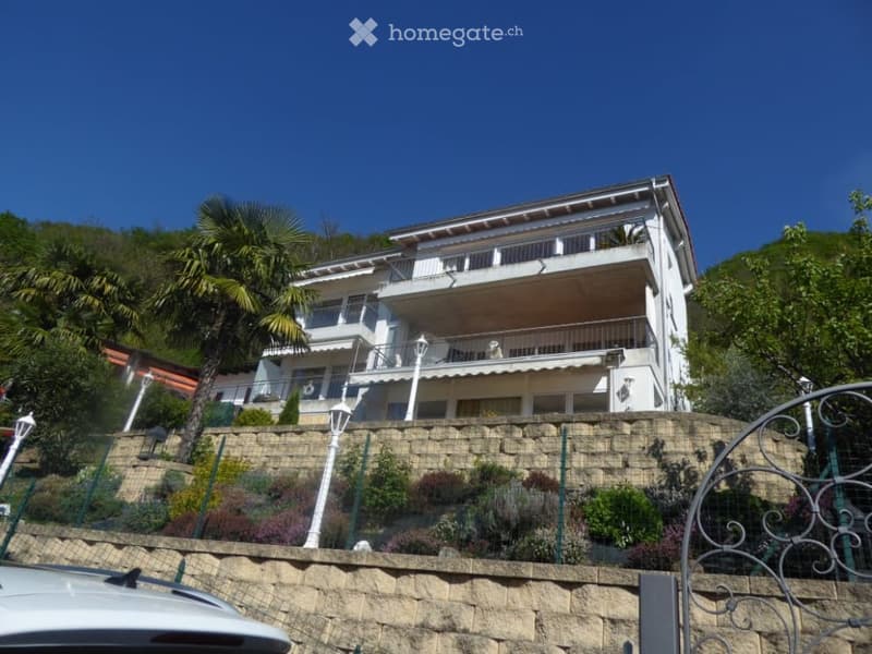 3-story apartment building with balconies, surrounded by palm trees and landscaping, with a stone retaining wall in the foreground