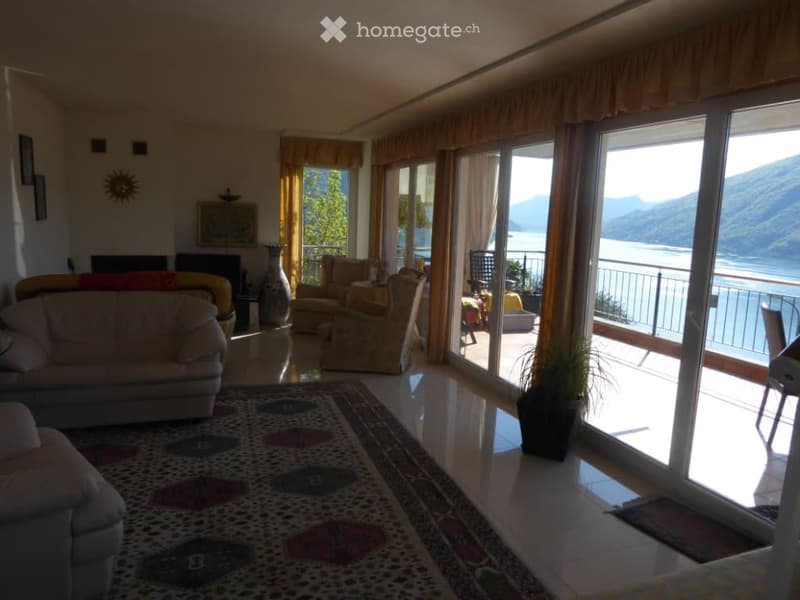 Spacious living room with large windows and sliding glass doors leading to a balcony with a scenic view of the lake and mountains. The room features comfortable sofas, a fireplace, and decorative elements like wall art and potted plants.