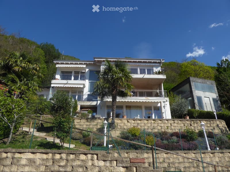 3-story white building with balconies, surrounded by palm trees and lush greenery, situated on a hillside with a stone retaining wall in the foreground