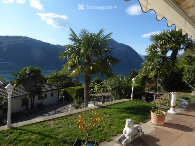 Lush green garden with palm trees, overlooking a lake or body of water surrounded by mountains in the background