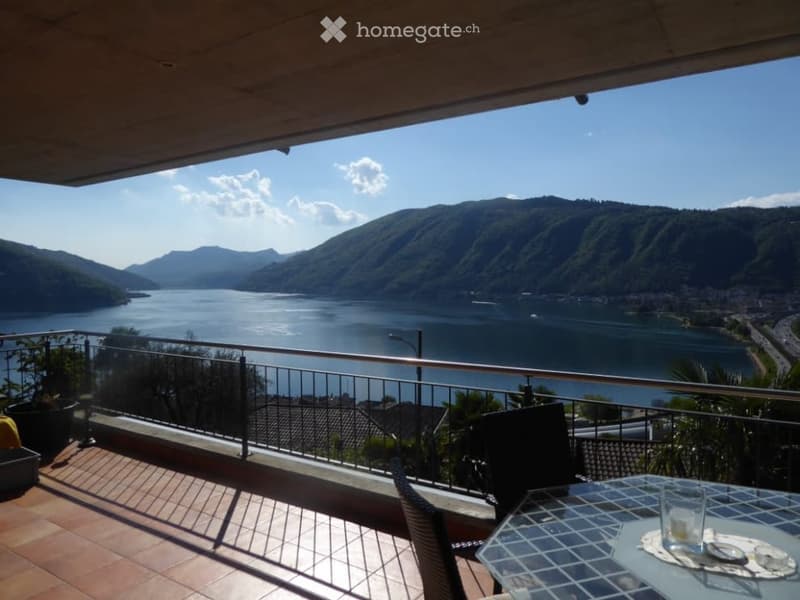 Balcony overlooking a scenic lake surrounded by mountains, with a table and chairs set up for outdoor dining.