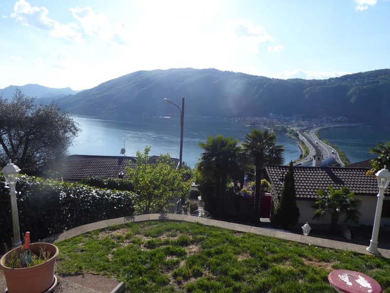 The image shows a scenic view of a lake surrounded by mountains and lush greenery. There are palm trees and other vegetation in the foreground, and a road can be seen in the distance. The view appears to be from a balcony or terrace overlooking the lake a
