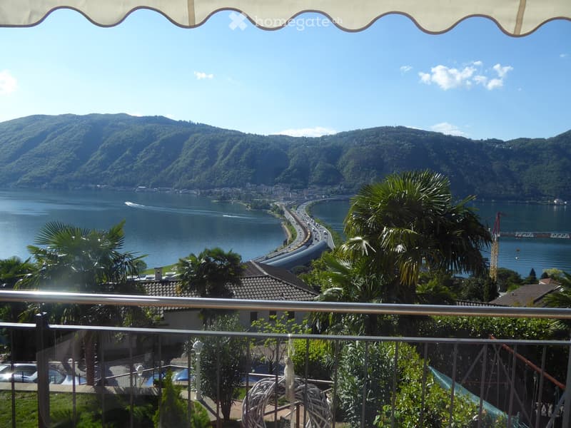 Scenic view of a lake surrounded by mountains, with a winding road along the shoreline. The balcony overlooks lush greenery and palm trees.