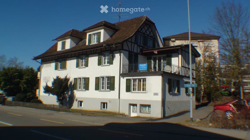 A two-story residential building with white walls, green shutters, and a brown roof. Features include multiple windows, a balcony on the upper floor, and a signboard on the balcony.