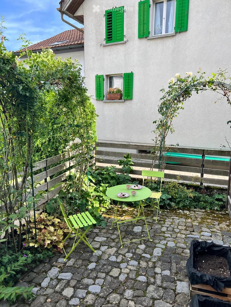A small garden area with a green metal table and chairs, surrounded by plants and a wooden fence, with a house in the background featuring green shutters and a window box.