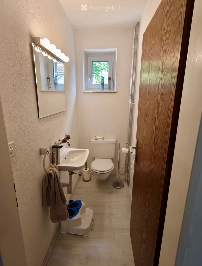 Compact bathroom with a white sink, toilet, towel, mirror, and a small window. Includes a step stool and a light fixture above the mirror.