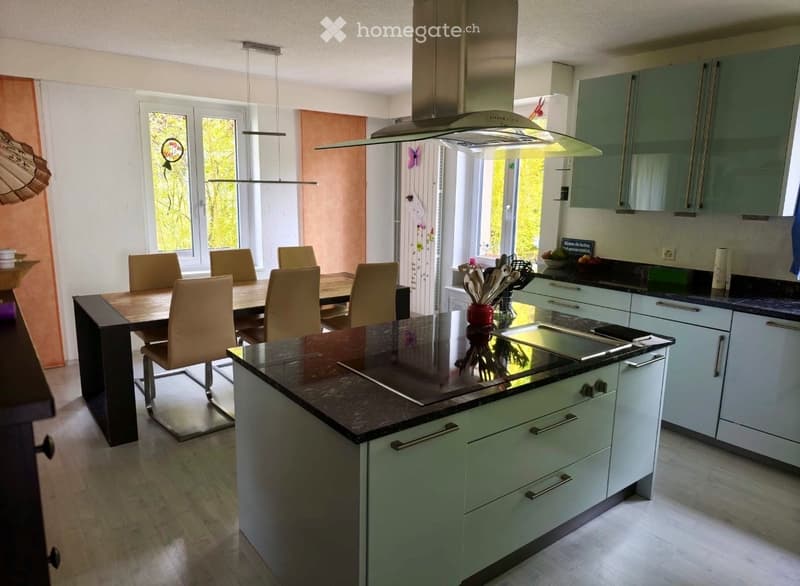 Modern kitchen with light green cabinets, dark granite countertops, a central island, and a dining area with a wooden table and beige chairs. Features include an induction cooktop, range hood, and pendant lighting.