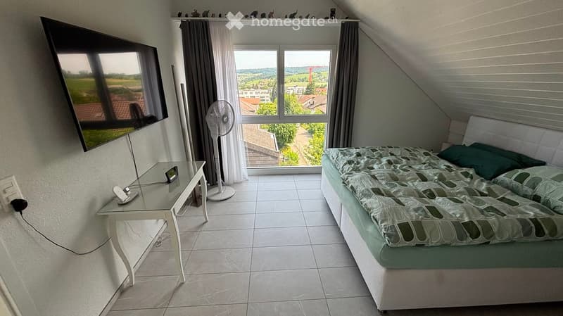 Bright bedroom with a bed, a small table, a fan, and a large window with a scenic view.
