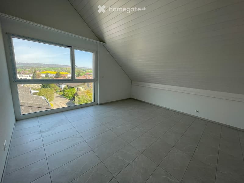 An empty room with a large window, wooden ceiling, and tiled floor. The room has a view of the outside with houses and trees.