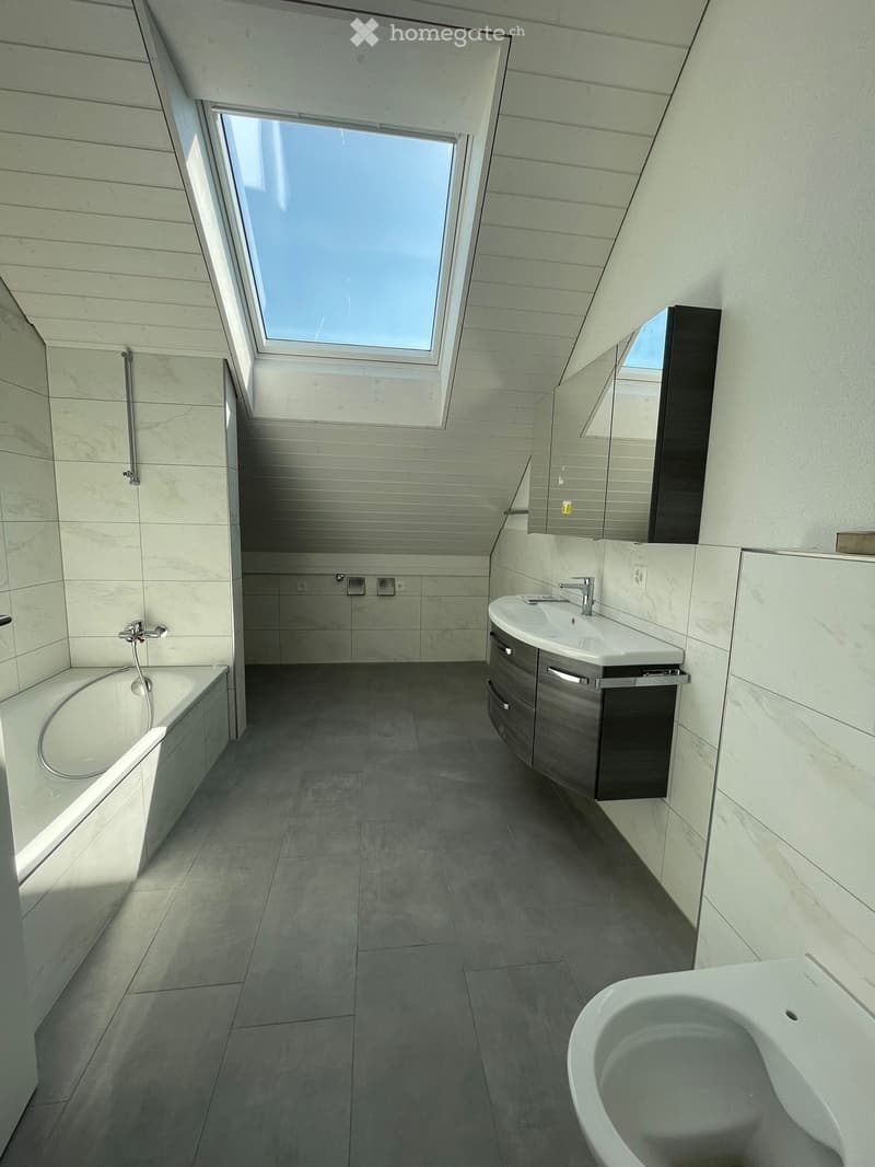 Modern bathroom with white tiles, bathtub, sink, and toilet. Features a skylight and a mirror above the sink.