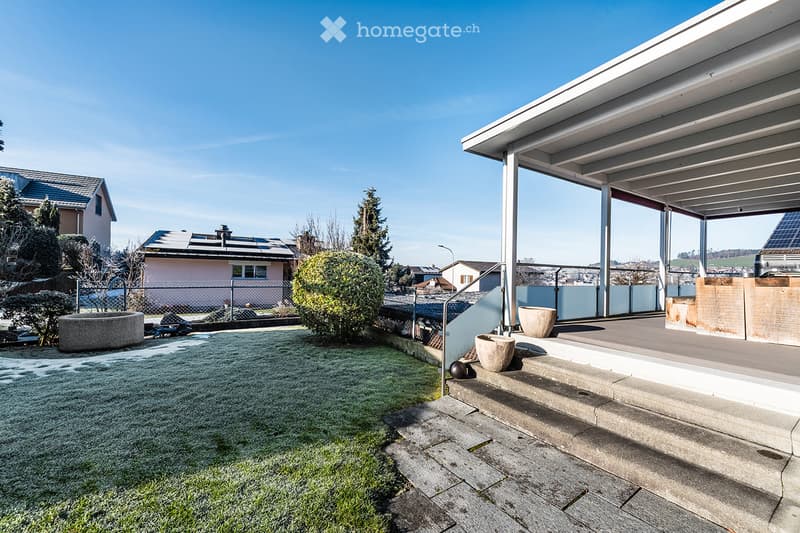 A modern outdoor area with a covered patio, steps leading up, decorative pots, and a small lawn area.