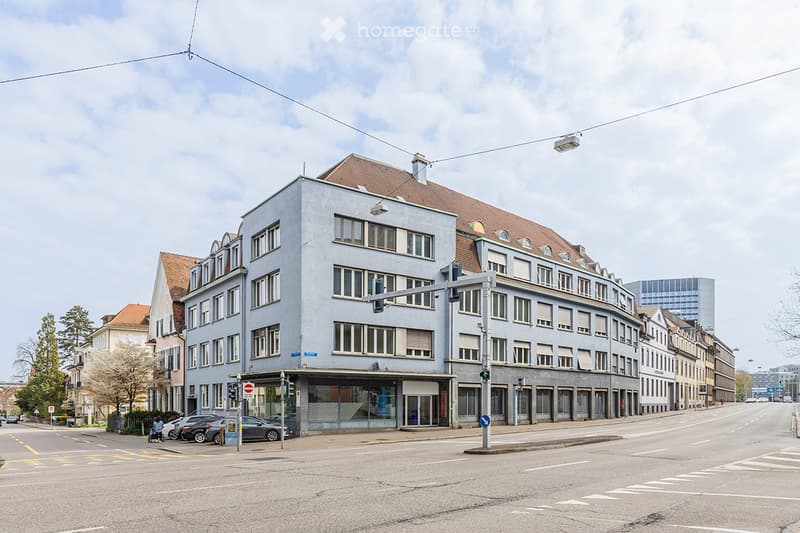 Multi-story building with light blue facade, multiple windows, and a curved corner section. The building is situated at a street corner with traffic lights and a few parked cars visible.