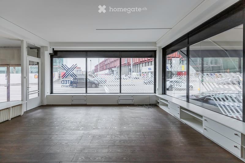 Empty room with large windows, wooden floor, white built-in storage units, and a long white bench along one wall.