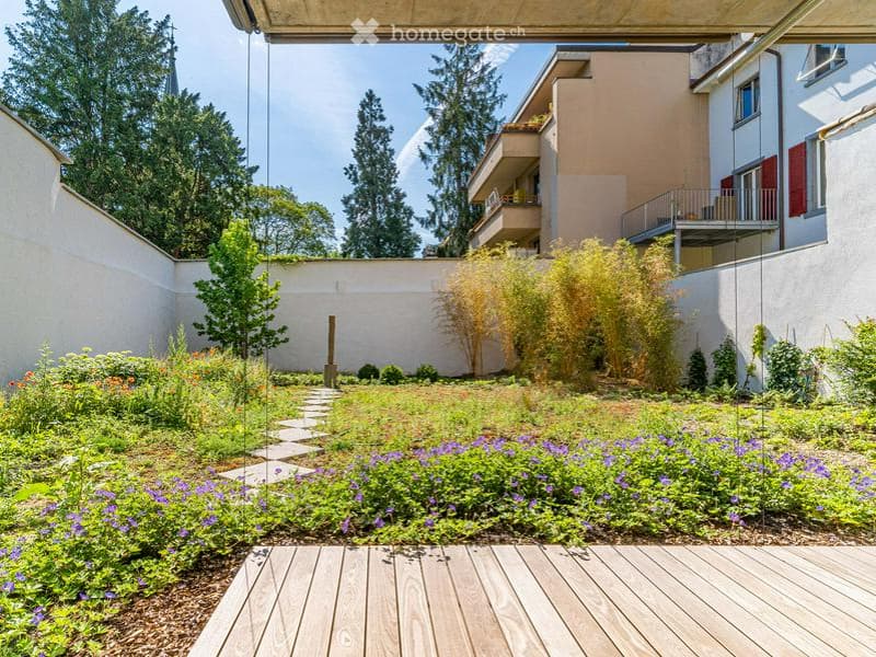 Well-maintained garden with a winding stone pathway, colorful flowering plants, shrubs, and trees, surrounded by modern apartment buildings.