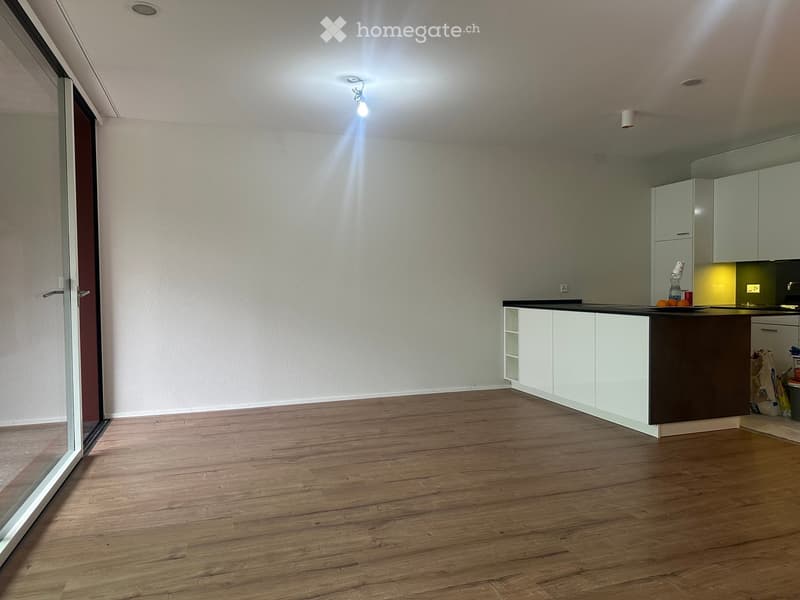 Empty kitchen with white cabinets, dark countertop, wooden flooring, and a large window with blinds.