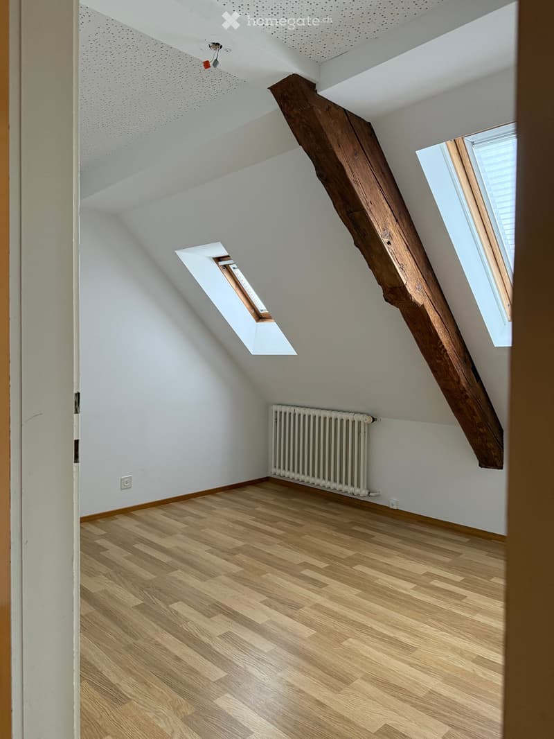 Empty attic room with wooden floor, white walls, exposed wooden beam, skylight window, and radiator.