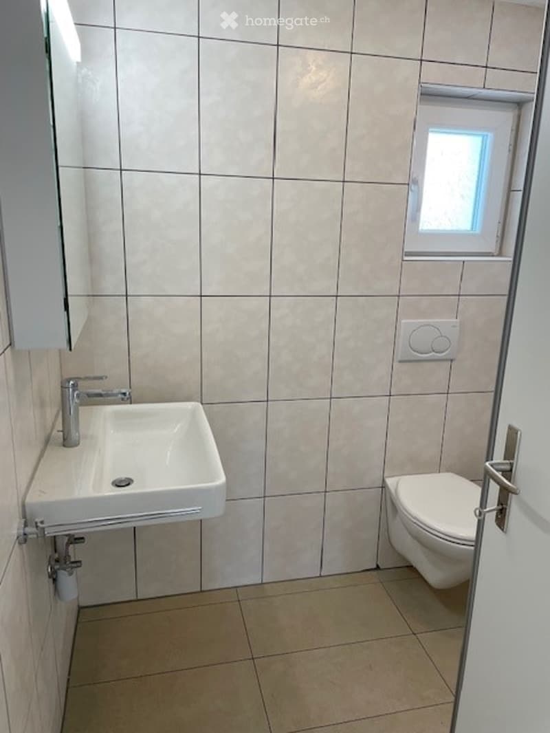 Modern bathroom with beige tiled walls and floor, a white sink with a chrome faucet, a toilet, a mirror cabinet, and a small window for natural light.