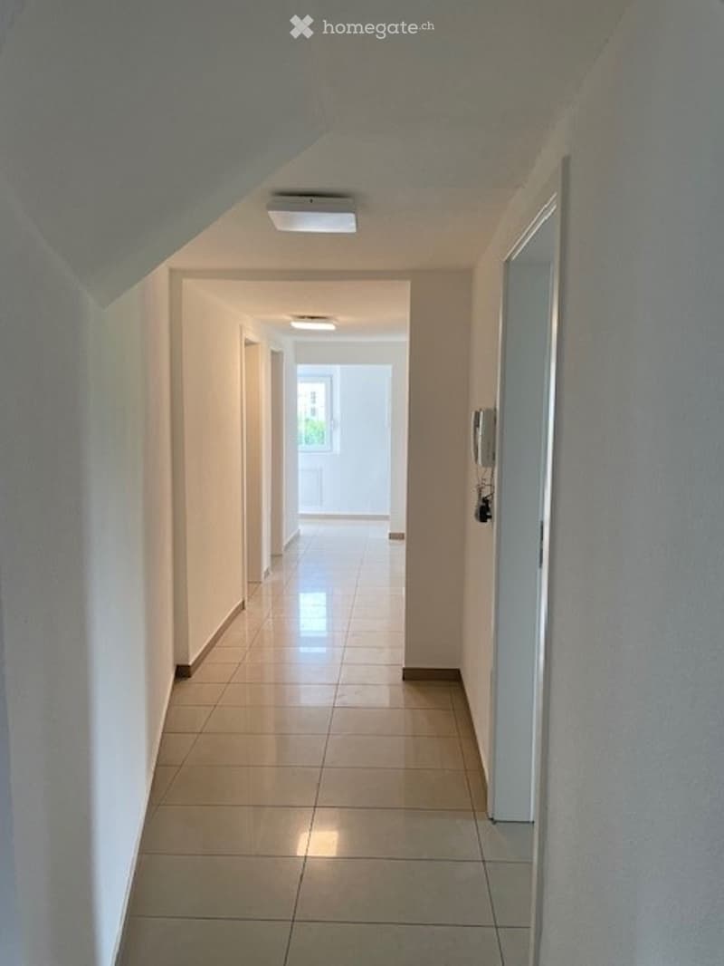 A clean, well-lit hallway with tiled flooring and white walls, featuring recessed lighting and a partially open door at the end.