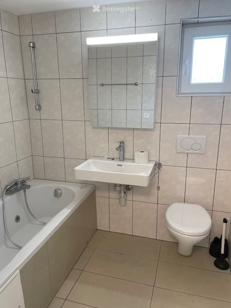 Modern bathroom with beige tiled walls and floor, featuring a bathtub, sink, toilet, and a large mirror with built-in lighting above the sink.