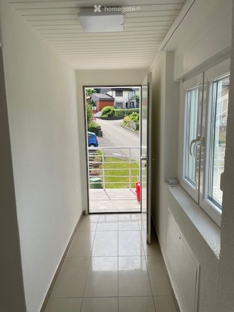 Bright hallway with white walls, tiled floor, and a large window on the right side. An open door at the end leads to an outdoor area with a view of a street and parked car.