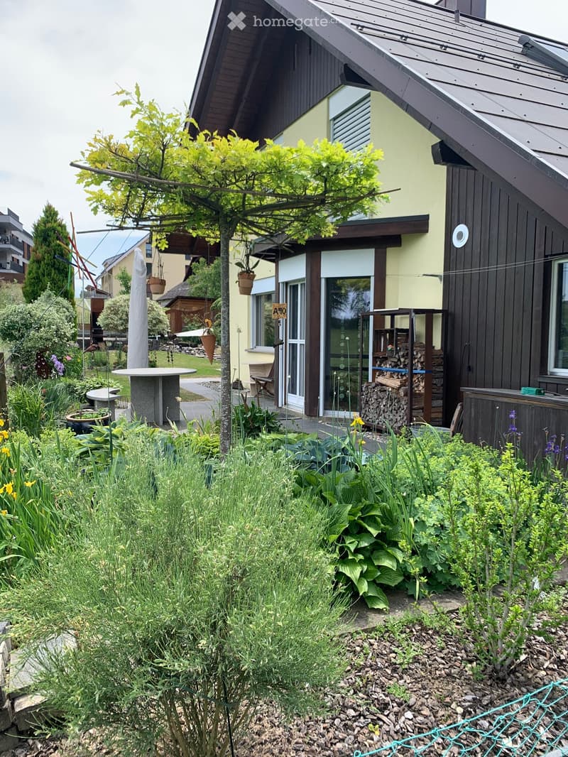 Garden area with a modern house in the background, featuring a variety of plants, a round table, a stack of firewood, and a hanging basket.