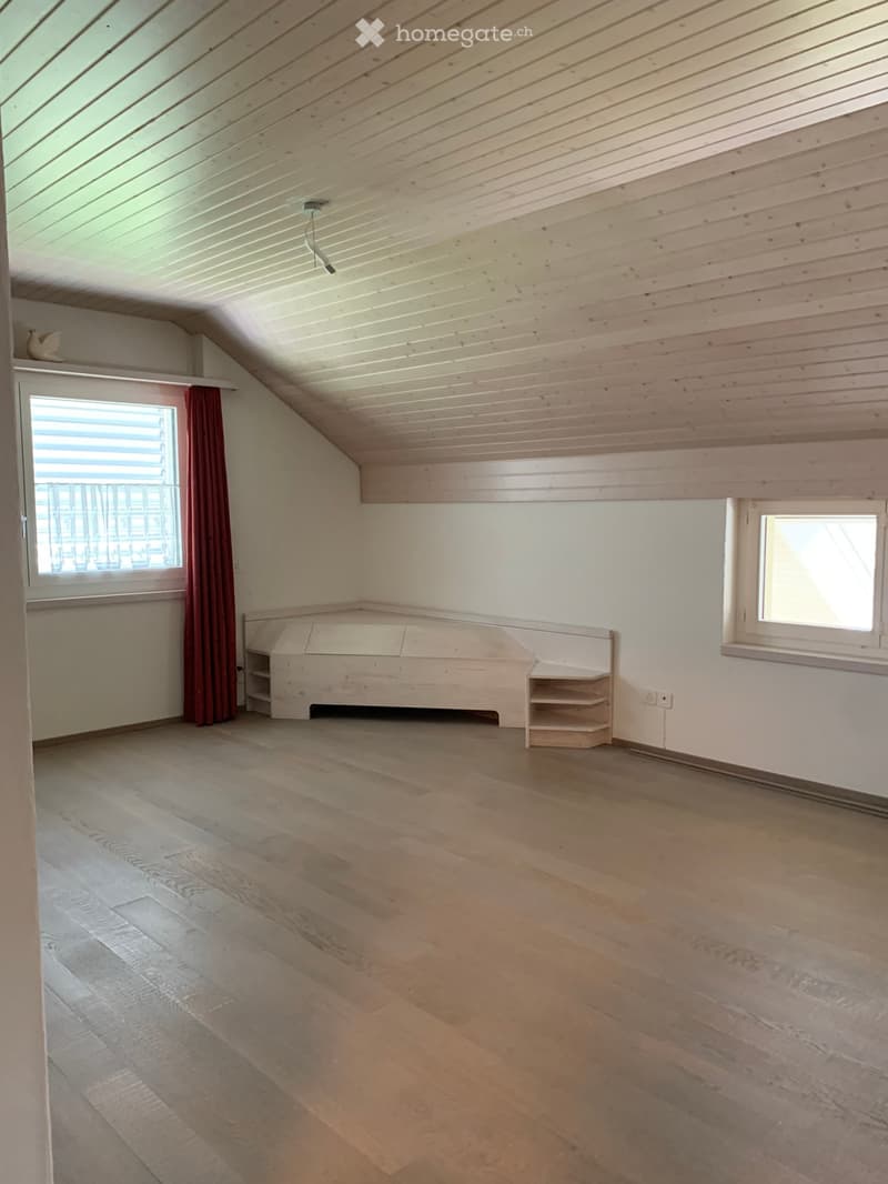 Empty attic room with wooden floors, white walls, and exposed wooden ceiling beams. Features two windows, a built-in bench, and a small shelf unit.