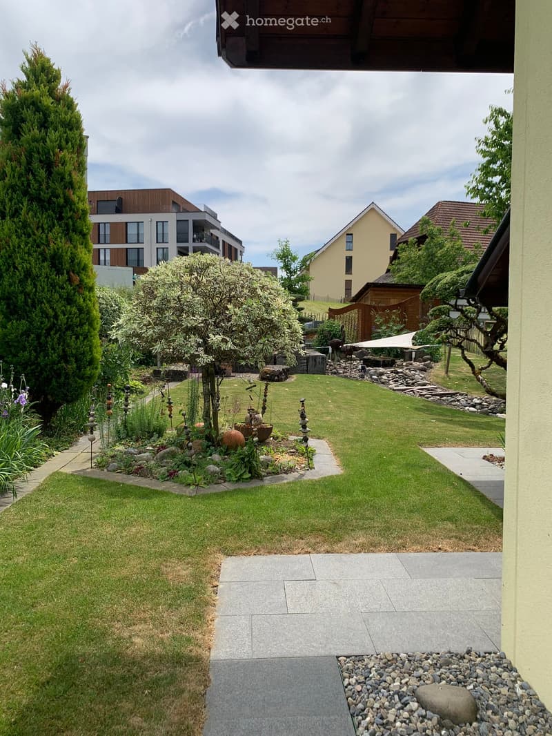 Well-maintained garden with a lawn, stone pathways, decorative rocks, and surrounding trees and shrubs.