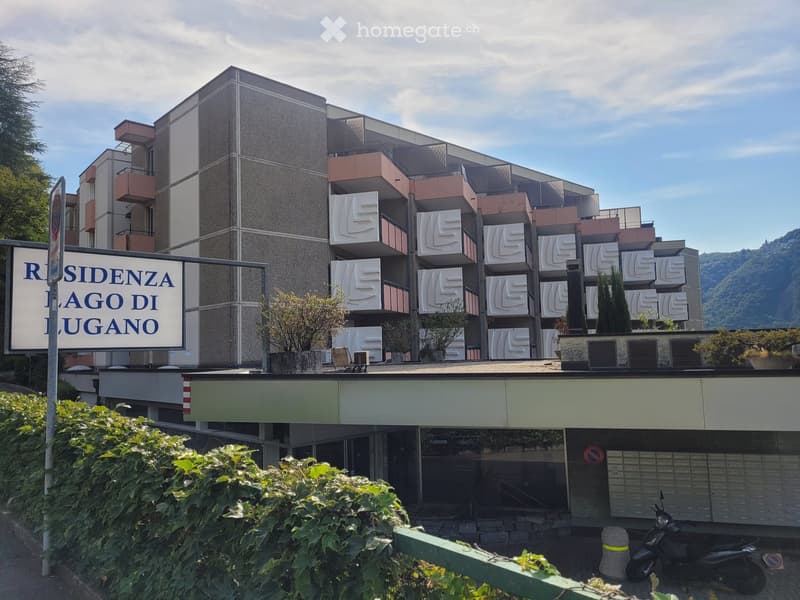 Modern multi-story residential building with balconies and geometric facade design, featuring a sign reading "RESIDENZA LAGO DI LUGANO".
