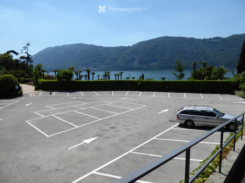 Parking lot with marked spaces, a single parked car, surrounded by greenery and a scenic lake and mountain view.