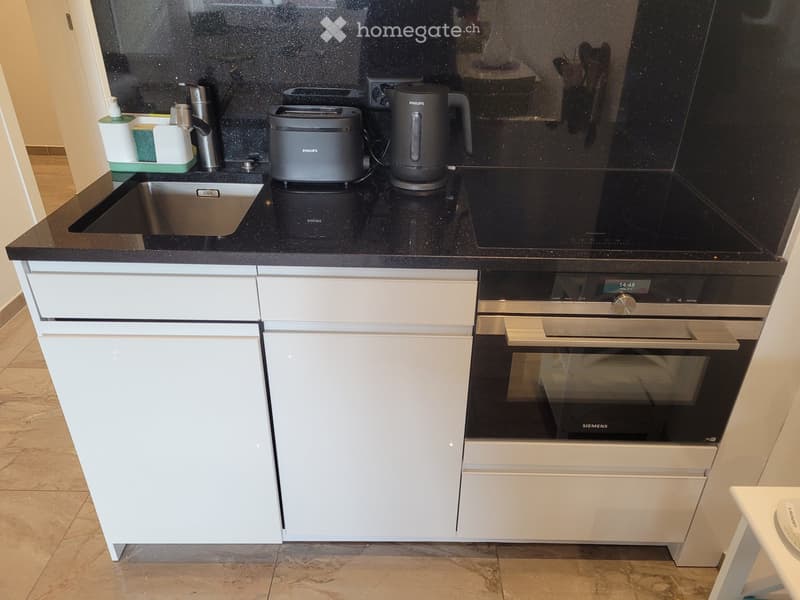 Modern kitchen with white cabinets, black countertops, stainless steel sink, and built-in oven. Includes a toaster and electric kettle on the counter.