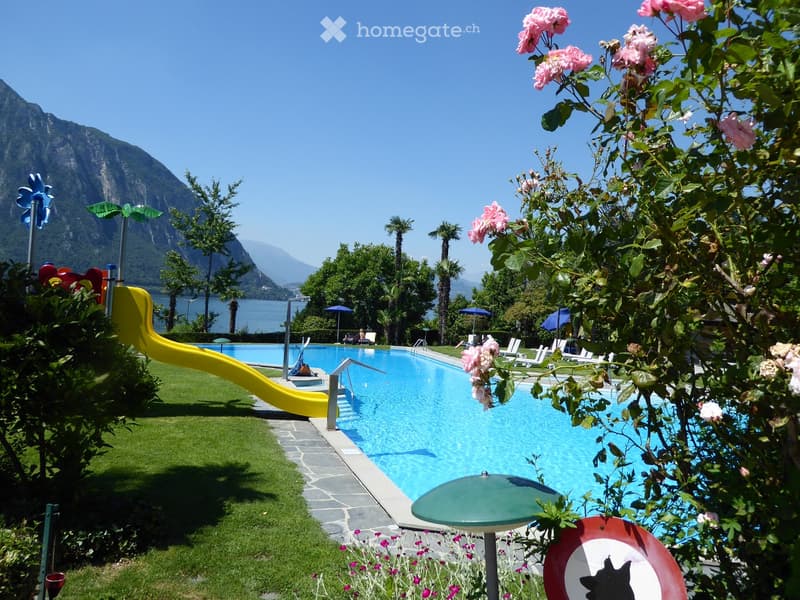 Outdoor area with a swimming pool, surrounded by greenery, a yellow slide, lounge chairs, and umbrellas, with a scenic mountain and lake view in the background.
