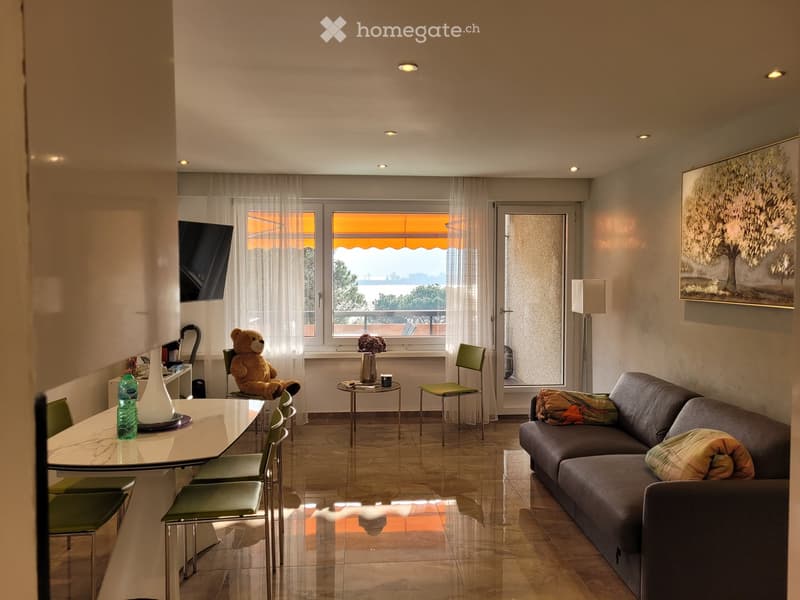 Bright living room with a view, featuring a sofa, coffee table, chairs, a lamp, a TV screen, and decorative items.