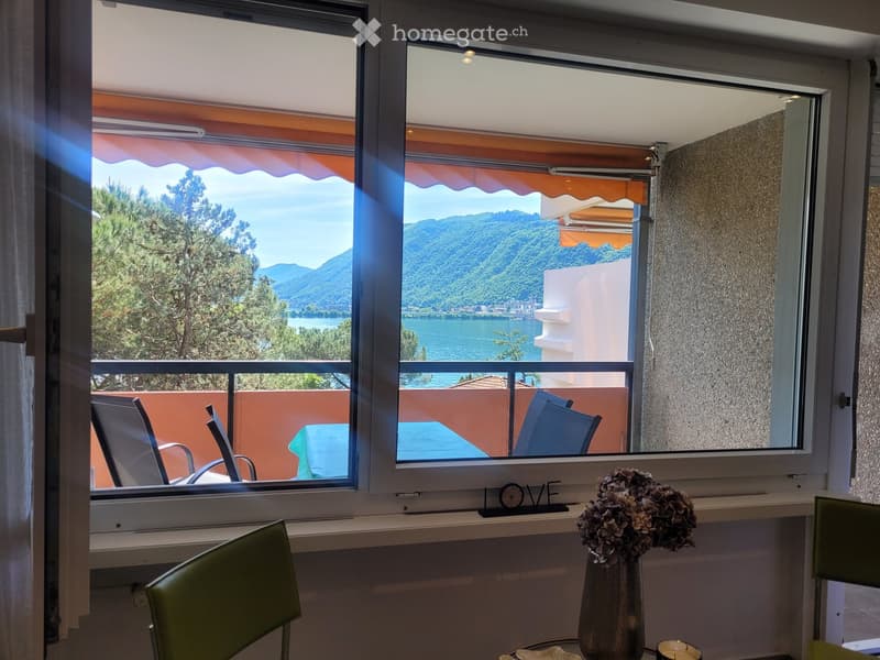 A furnished room with a view of a lake and mountains through large windows. The room features a green chair, a small table with a vase of flowers, and a decorative "LOVE" sign on the windowsill.