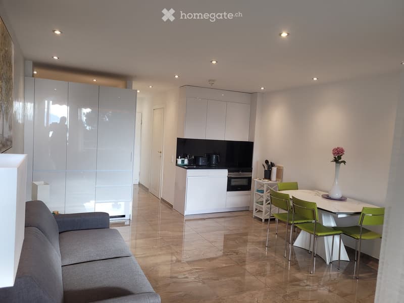 Modern open-plan living and dining area with a grey sofa, white dining table with green chairs, white cabinets, and a small kitchen area with an oven and countertop.