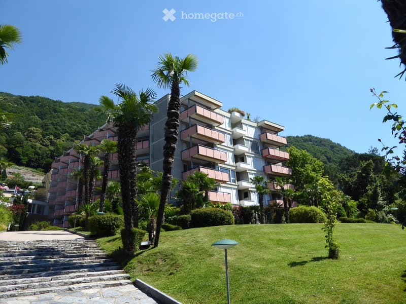 Modern multi-story residential building with pink balconies, surrounded by palm trees and a well-maintained lawn.