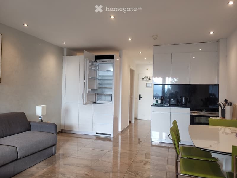 Modern open-plan living and dining area with a grey sofa, dining table with green chairs, white kitchen cabinets, refrigerator, and oven.
