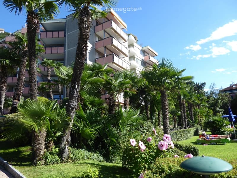 A well-maintained garden area with palm trees, flowering shrubs, and a multi-story building with balconies in the background.