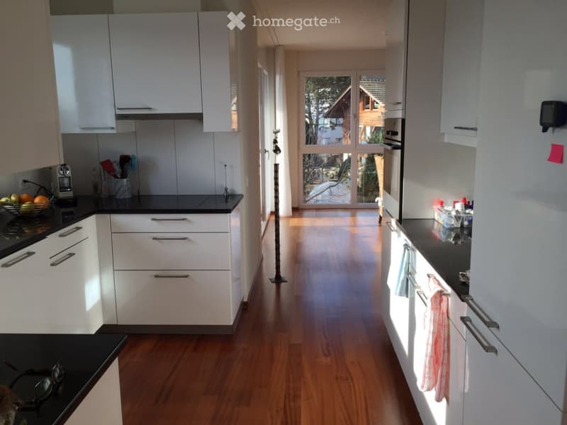 Modern kitchen with white cabinets, black countertops, and wooden flooring. Features a coffee maker, fridge, oven, and a view through large windows.