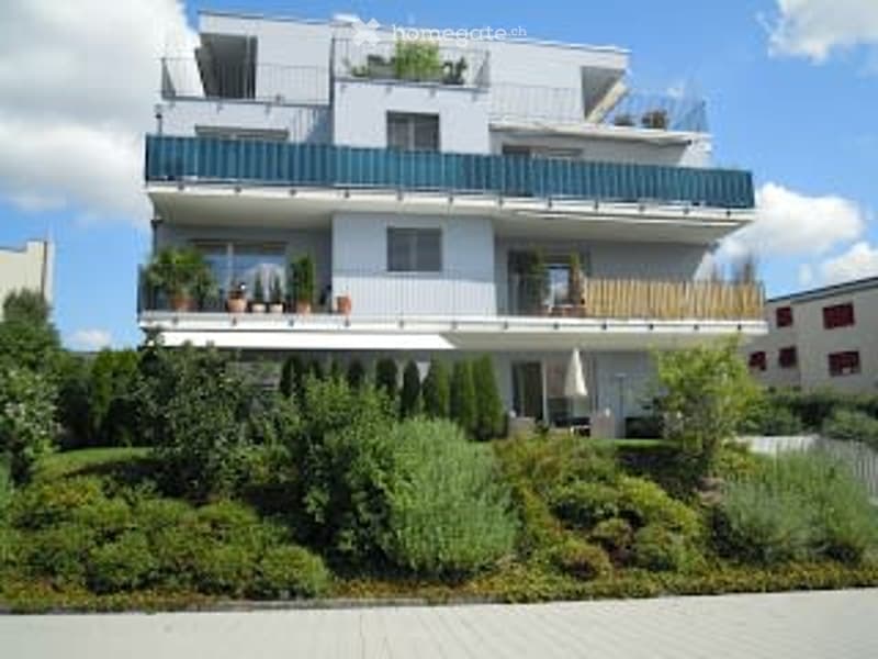 Modern multi-story apartment building with balconies, greenery, and a raised ground floor.