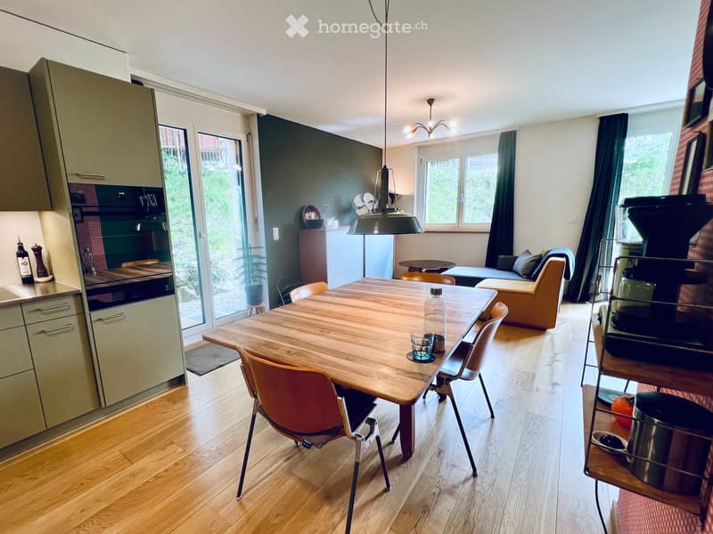 Open-plan living and dining area with wooden flooring, a large dining table with chairs, a sofa, and a modern kitchen area with cabinets and appliances.