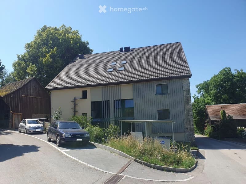Two-story house with a tiled roof, multiple windows, and a covered entrance area. Two cars are parked in front of the house, and there is a small garden area with plants and shrubs near the entrance.