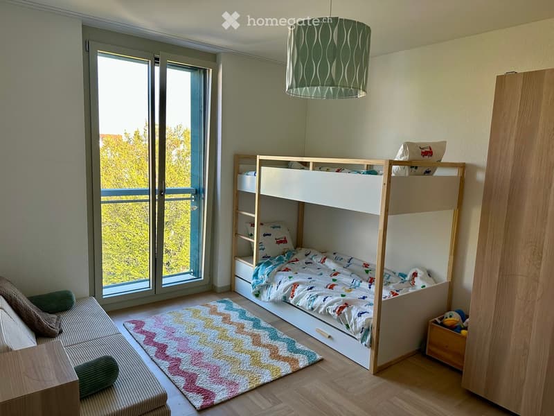 Bright bedroom with a bunk bed, colorful bedding, a sofa, a rug, and a view of greenery through large windows.