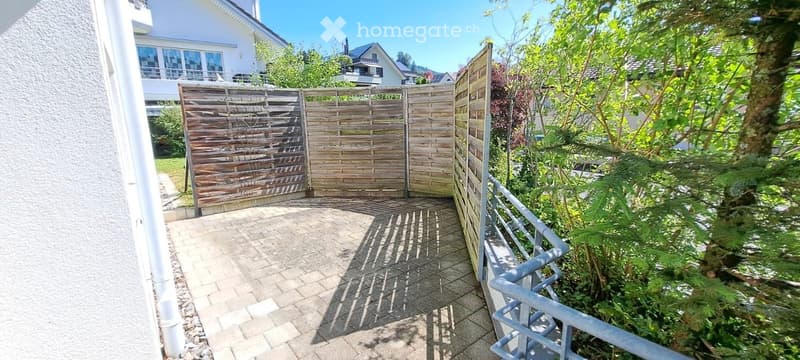 A paved outdoor area with a wooden gate, surrounded by greenery and a metal railing on one side.