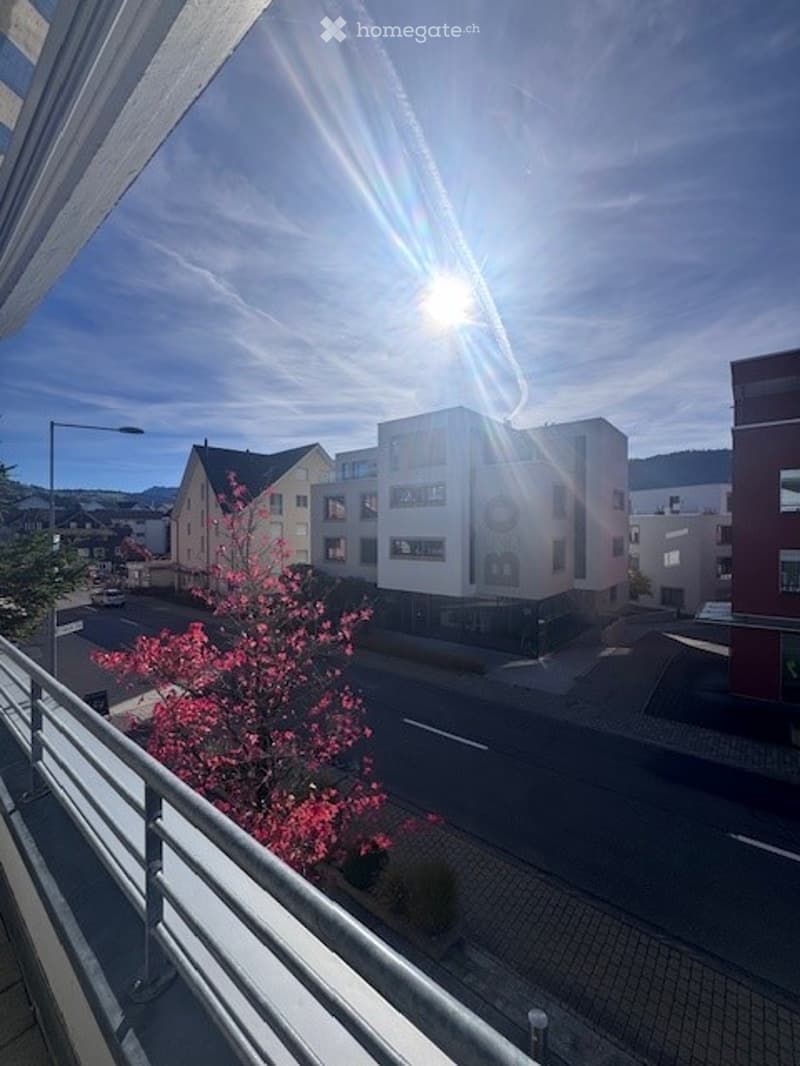 View from the balcony of a multi-storey residential building, with buildings, greenery, a road, and a bright sun in the sky
